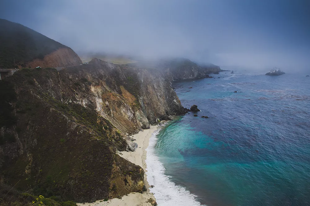 Photographie artistique des falaises escarpées de Big Sur en Californie avec plage isolée et eaux turquoise de l'océan Pacifique sous un ciel brumeux, tirage d'art paysage marin