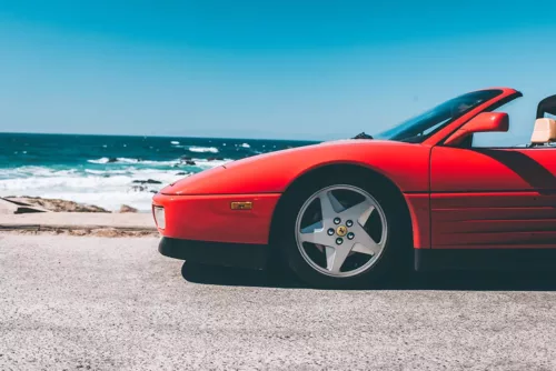 Photographie Ferrari Testa Rossa Spider Photographie artistique d'une Ferrari Testarossa Spider rouge stationnée au bord de l'océan Pacifique en Californie, avec vagues et rochers en arrière-plan sous un ciel bleu azur, tirage d'art automobile limité