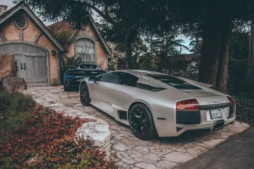 Photographie Lamborghini et McLaren Photographie artistique d'une Lamborghini argentée et d'une McLaren bleue devant un cottage rustique, contraste entre supercars modernes et architecture traditionnelle, tirage d'art automobile de luxe