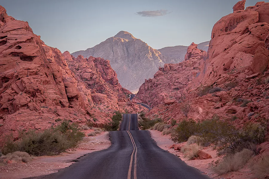 Photographie artistique de la route sinueuse traversant les formations rocheuses rouges de la Valley of Fire au Nevada, avec montagnes majestueuses au coucher du soleil
