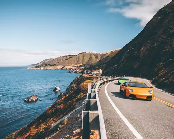 Photographie artistique de deux Lamborghini Murcielago LP 640 roulant sur la Highway One californienne avec vue spectaculaire sur l'océan Pacifique et les falaises côtières