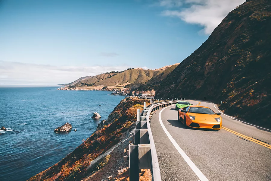 Photographie artistique de deux Lamborghini Murcielago LP 640 roulant sur la Highway One californienne avec vue spectaculaire sur l'océan Pacifique et les falaises côtières