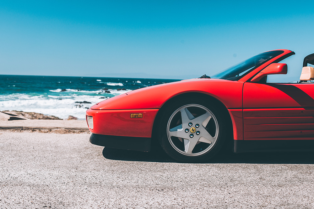 Photographie artistique d'une Ferrari Testarossa Spider rouge stationnée au bord de l'océan Pacifique en Californie, avec vagues et rochers en arrière-plan sous un ciel bleu azur, tirage d'art automobile limité
