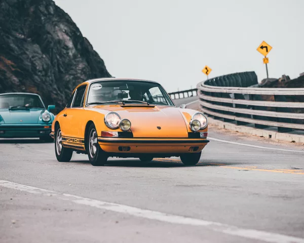 Photographie déco murale de Porsche 911 classique orange sur route côtière sinueuse suivie d'une Porsche bleu turquoise, parfaite pour décoration intérieure automobile vintage