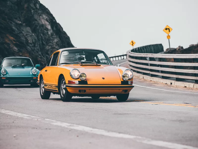 Photographie déco murale de Porsche 911 classique orange sur route côtière sinueuse suivie d'une Porsche bleu turquoise, parfaite pour décoration intérieure automobile vintage