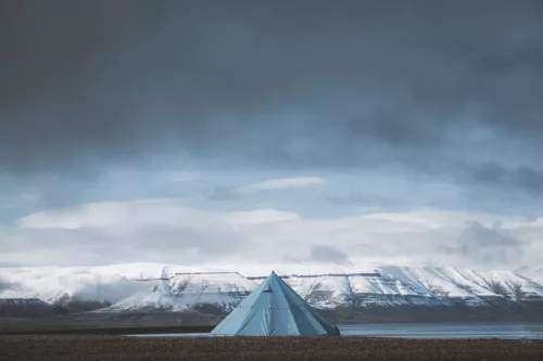 Photographie de paysage du Svalbard montrant une tente tipi bleue solitaire face aux montagnes enneigées des fjords arctiques sous un ciel dramatique