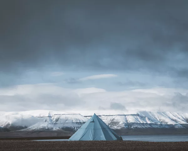 Photographie de paysage du Svalbard montrant une tente tipi bleue solitaire face aux montagnes enneigées des fjords arctiques sous un ciel dramatique