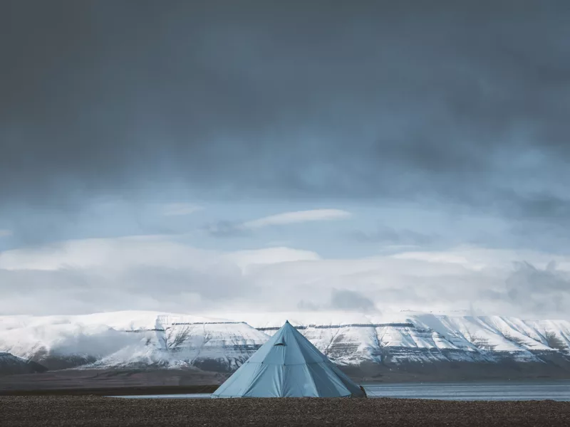 Photographie de paysage du Svalbard montrant une tente tipi bleue solitaire face aux montagnes enneigées des fjords arctiques sous un ciel dramatique
