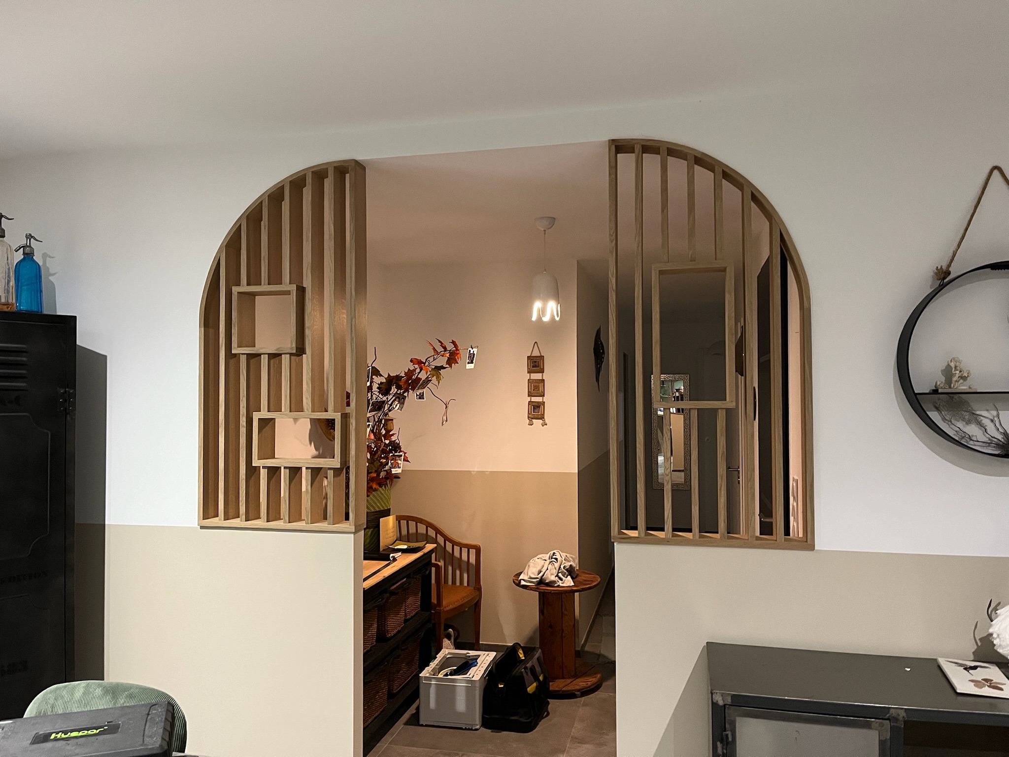 Arche en bois sculpté divisant un espace intérieur, avec étagères et poutres verticales, décor floral et chaise en bois, ambiance chaleureuse et moderne. Arche en bois sculpté divisant un espace intérieur, avec étagères et poutres verticales, décor floral et chaise en bois, ambiance chaleureuse et moderne.