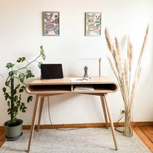 Bureau moderne en bois clair avec tablette incurvée, ordinateur portable, pot de plantes à gauche, pots de stylos et dessins sur le bureau, assise blanche, rangée de tiges deherbe au droit, et posters japonais au mur.