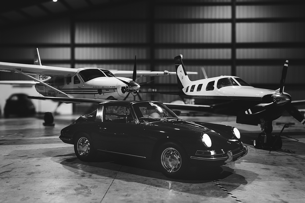 Voiture sportive classique noire au premier plan dans un hangar, entourée d’avions privés blancs, plancher en béton et éclairage industriel en arrière-plan. Voiture sportive classique noire au premier plan dans un hangar, entourée d’avions privés blancs, plancher en béton et éclairage industriel en arrière-plan.