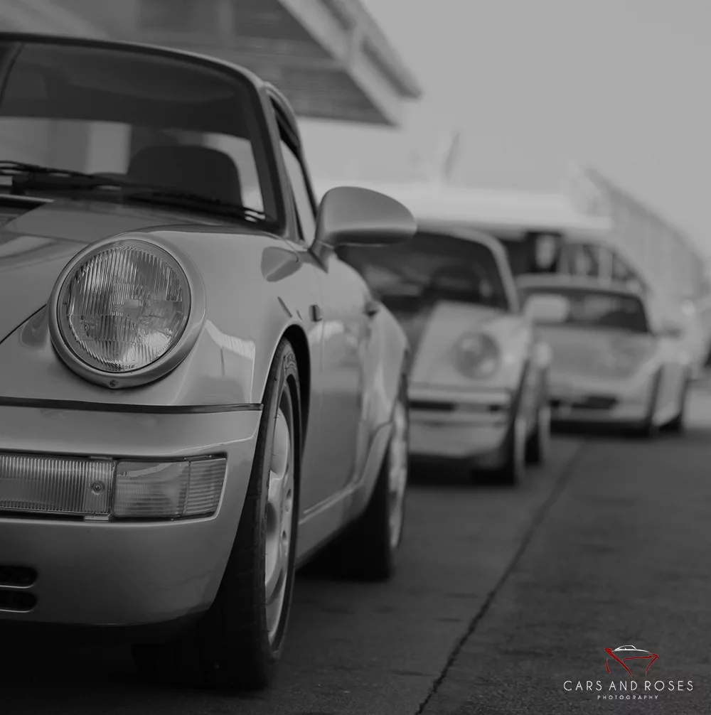 Voitures de sport classiques alignées en rang, premier coupé Porsche gris en gros plan, flou vers l’arrière, logo Cars and Roses en bas à droite.