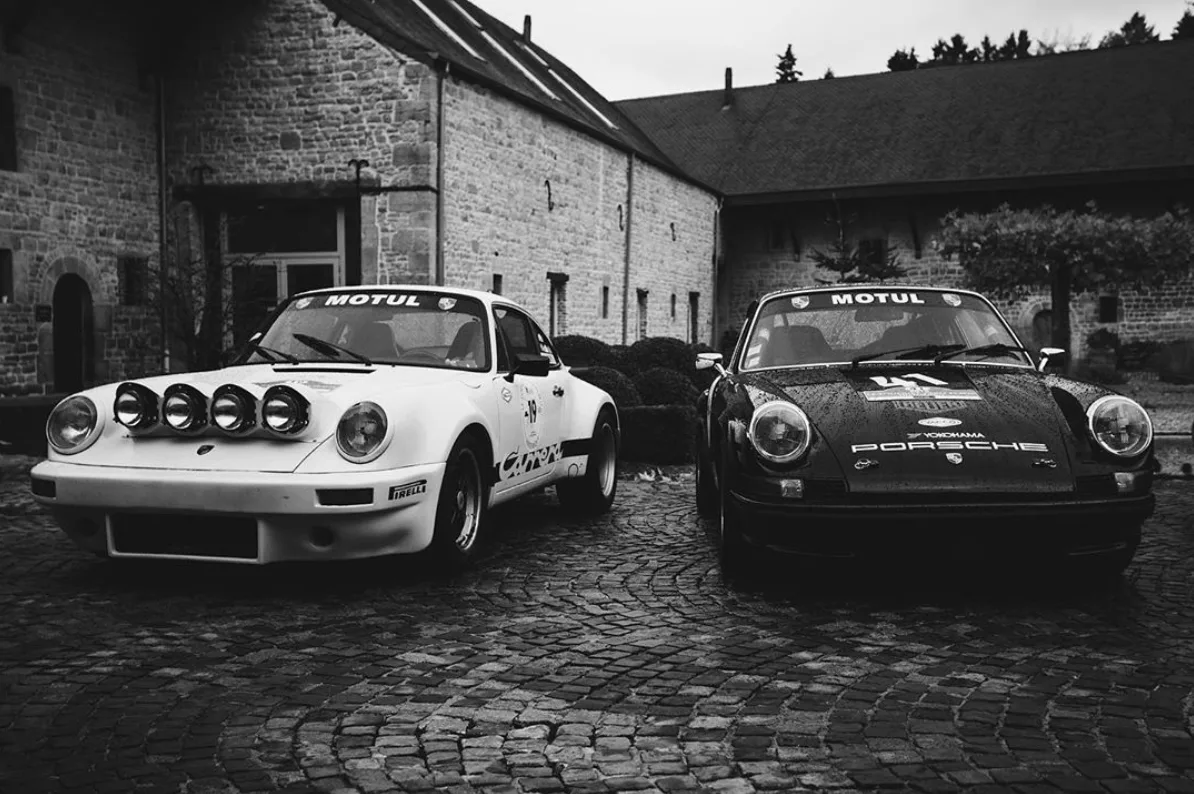 Deux Porsche de rallye vintage garées sur une ruelle pavée devant un bâtiment en pierre, vue en noir et blanc.