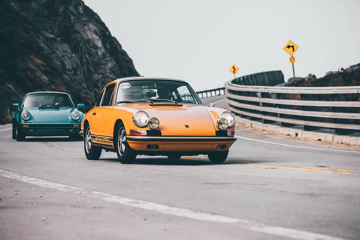 Deux Porsche anciennes en conduite sur une route côtière, l’une jaune au premier plan et l’autre bleue en arrière-plan, dans un virage près d’une rambarde. Comment photographier des voitures anciennes.