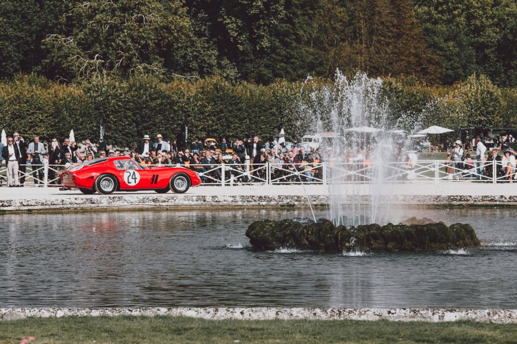 Ferrari 250 GTO survolant la rive d’un lac près d’une fontaine centrale, entourée d’un public et d’un écrin de verdure. Ferrari 250 GTO survolant la rive d’un lac près d’une fontaine centrale, entourée d’un public et d’un écrin de verdure.