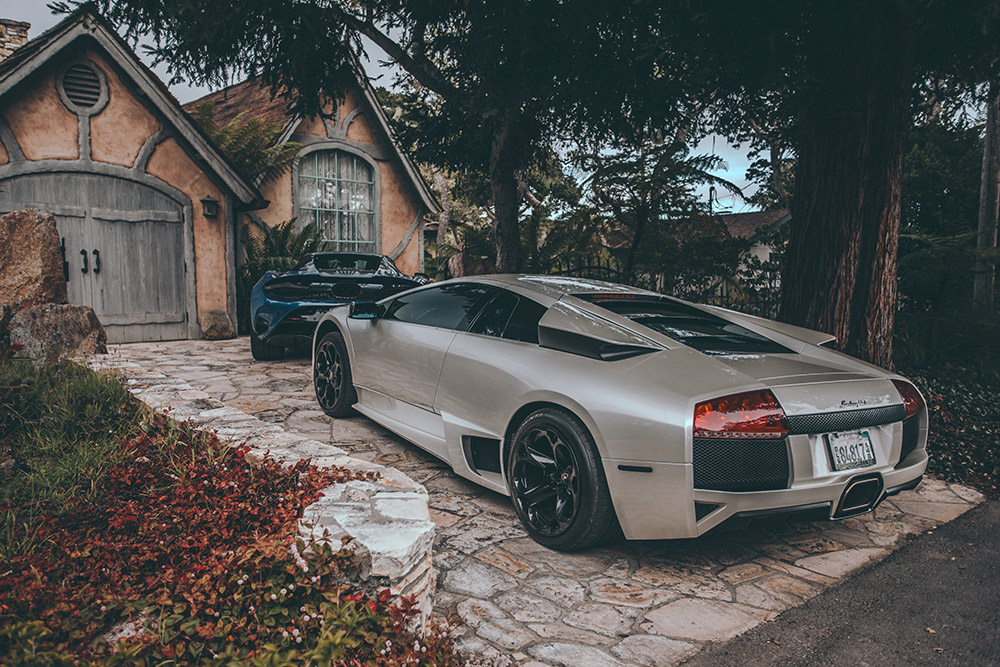 Lamborghini noir et argent garé sur une allée en pierres devant une maison de style cottage, entouré d’arbres et de verdure.