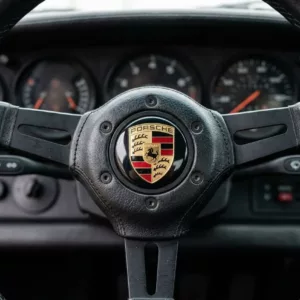 Volant noir avec écusson Porsche au centre, détails du tableau de bord en arrière-plan visible derrière. Pourquoi le logo porsche est un cheval cabré intégré naturellement.