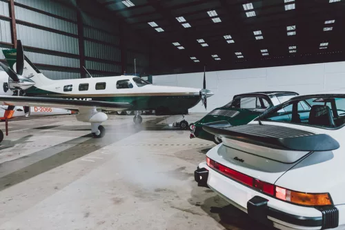 Porsche 911 Turbo and Piper Malibu, Hangar Light Porsche 911 Turbo blanche et 911 verte devant un Piper Malibu dans un hangar éclairé par des verrières - tableau photographique d'art
