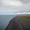 Vue d’en haut d’une plage de sable noir sur la côte islandaise, océan Atlantique gris, plaines vertes et montagnes enneigées