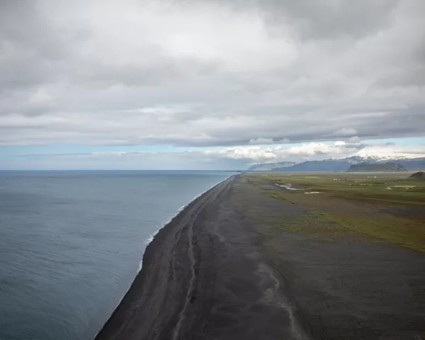 Photographie plage de sable noir Islande Vue d’en haut d’une plage de sable noir sur la côte islandaise, océan Atlantique gris, plaines vertes et montagnes enneigées