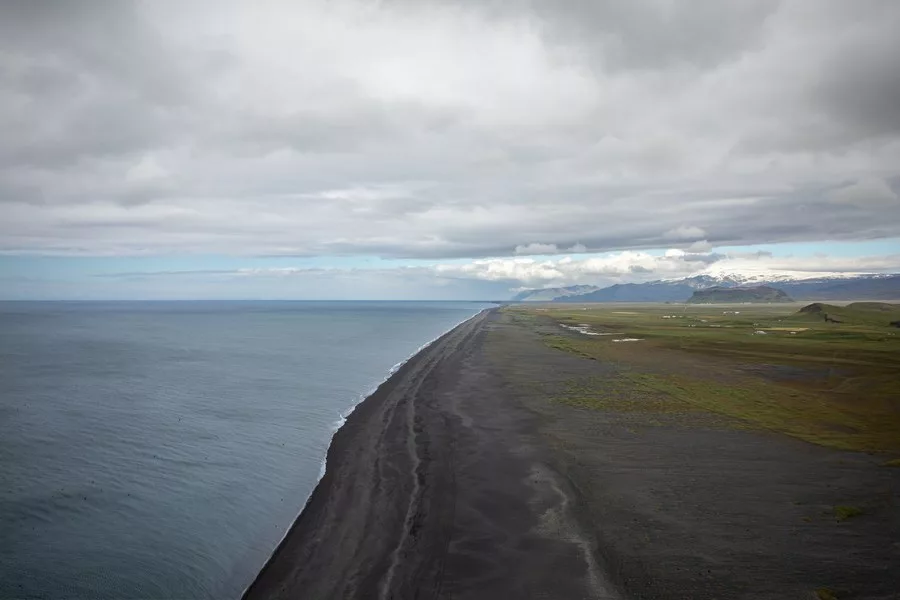 Vue d’en haut d’une plage de sable noir sur la côte islandaise, océan Atlantique gris, plaines vertes et montagnes enneigées