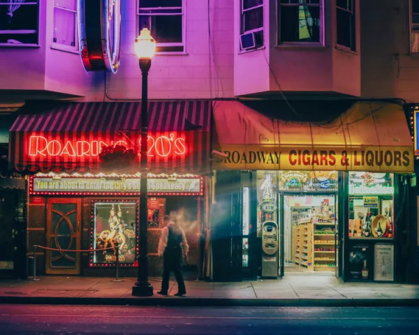 Photographie d’art de Broadway à San Francisco la nuit : enseigne rouge du Roaring 20’s, lampadaire allumé et passant en mouvement