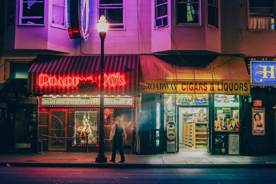 Photographie d’art de Broadway à San Francisco la nuit : enseigne rouge du Roaring 20’s, lampadaire allumé et passant en mouvement