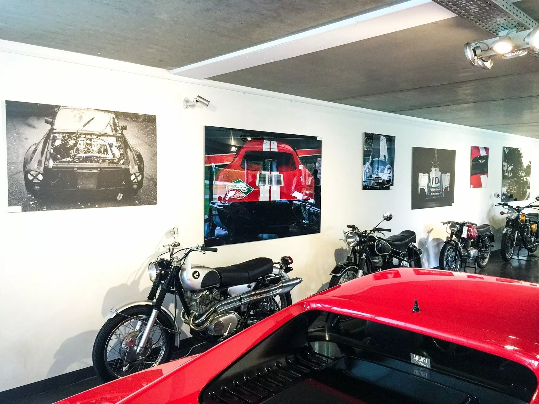 Tirages fine art sur aluminium et sous plexiglas accrochés au mur blanc d'un garage avec des motos anciennes et une voiture de sport rouge